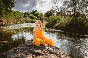 A joyful girl in a bright yellow outfit sits on a rock by a river, laughing with arms outstretched.