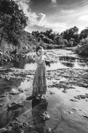 Black and white photo of a girl in a flowy dress standing on rocks in a shallow river surrounded by trees.