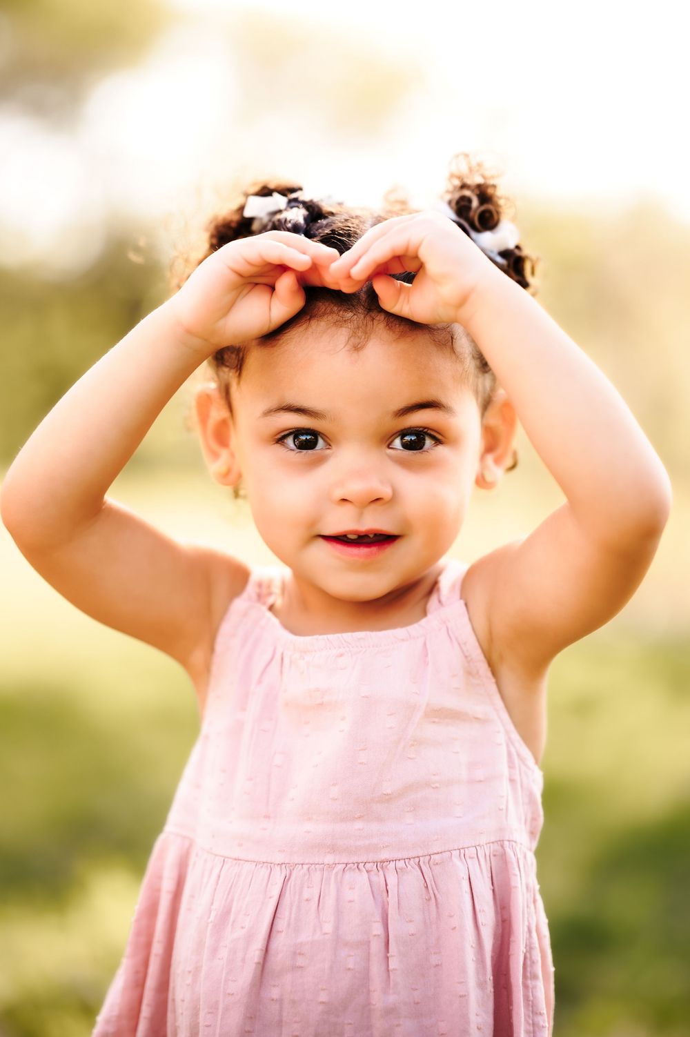 Adorable toddler girl in pink dress holding her curly pigtails outdoors in sunlight.