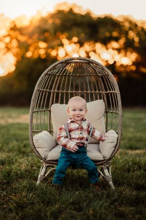Smiling baby boy in plaid shirt and suspenders sitting in a wicker egg chair outdoors at sunset.