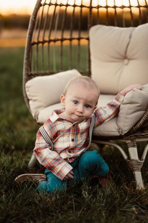 Adorable baby boy in plaid shirt and teal pants sitting beside a wicker egg chair on grass at golden hour.