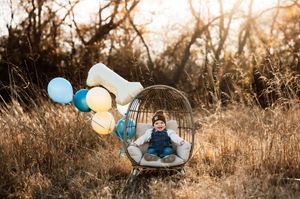 Happy baby boy sitting in a wicker chair with colorful balloons and number one in a field.