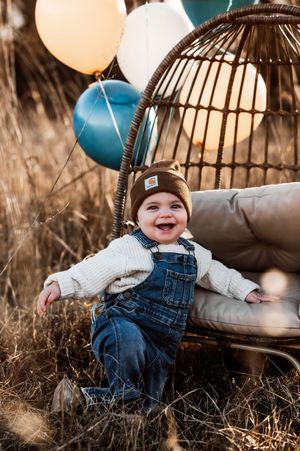 Laughing baby boy in Carhartt beanie and denim overalls sitting by balloons in autumn field.