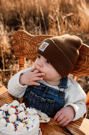 Adorable baby boy in Carhartt beanie and overalls eating birthday cake in a golden field.