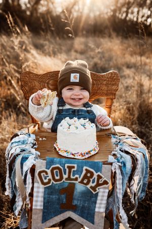 Smiling baby boy named Colby celebrating first birthday with white cake in a sunny field.