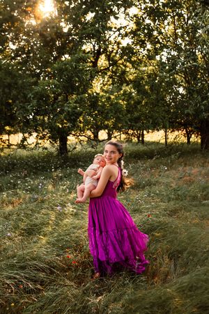 A smiling girl in a purple dress holds a baby while standing in a sunlit green meadow with trees.