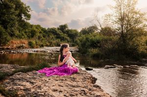 A girl in a purple dress sits on rocks by a river, gently holding a baby in a natural outdoor setting.