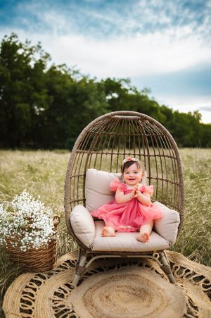 Happy toddler girl in pink dress sitting in wicker egg chair surrounded by wildflowers in a field.