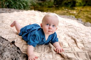 Blue-eyed baby in teal romper lying on knit blanket outdoors near water.
