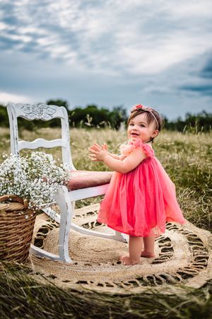 Baby girl in coral dress standing by white rocking chair in meadow.