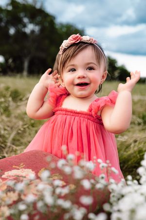 Smiling baby girl in coral dress and floral headband clapping hands outdoors in a wildflower meadow.