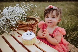 Baby girl in pink dress sitting beside cupcake and wildflowers outdoors.