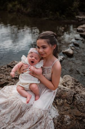 A young girl in a white lace dress sits on rocks by a river, cradling a yawning baby in her arms.