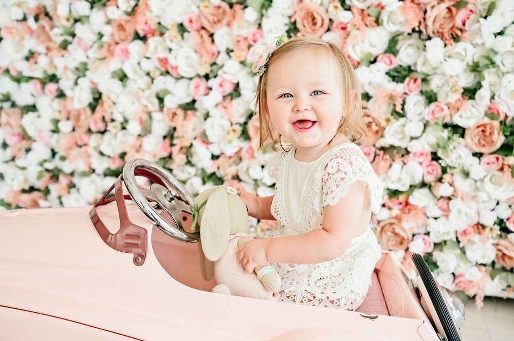 Happy baby girl in lace dress sitting in pink toy car against floral backdrop.