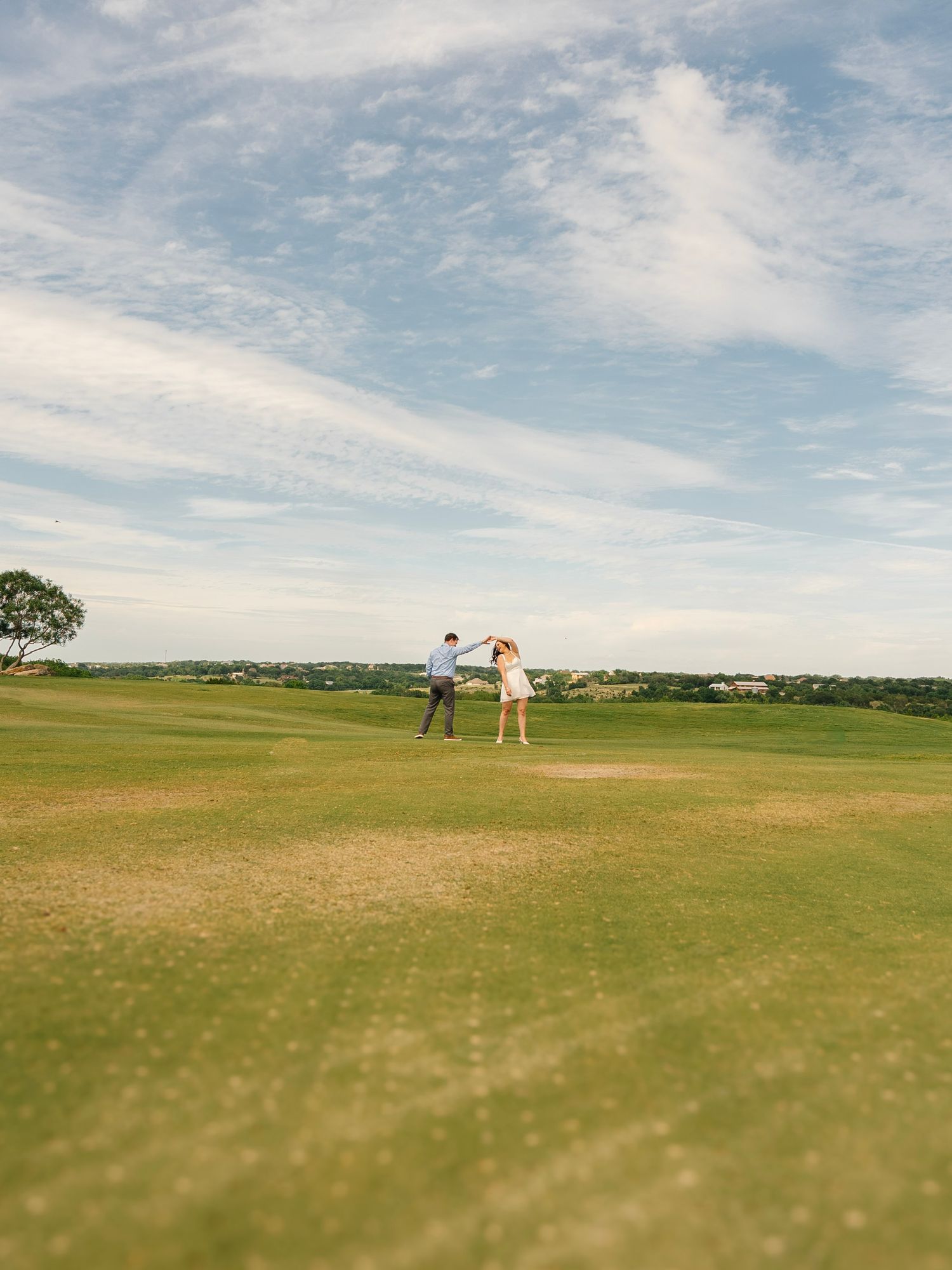 Couple holding hands on a lush green golf course under a wide blue sky with wispy clouds.
