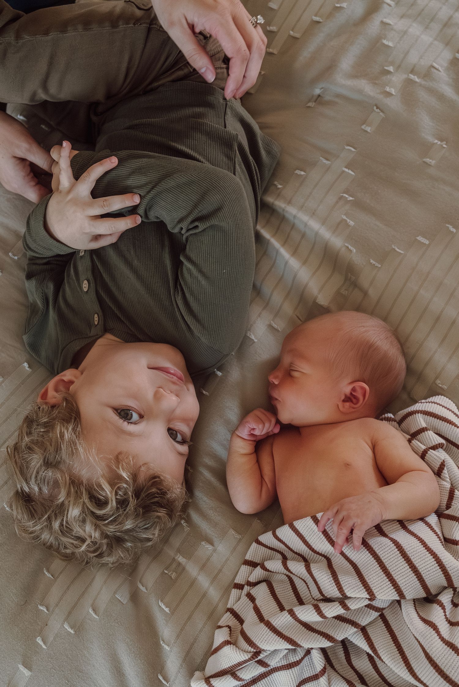 A young child in green lies beside a sleeping newborn baby on a beige textured bed with a striped blanket.