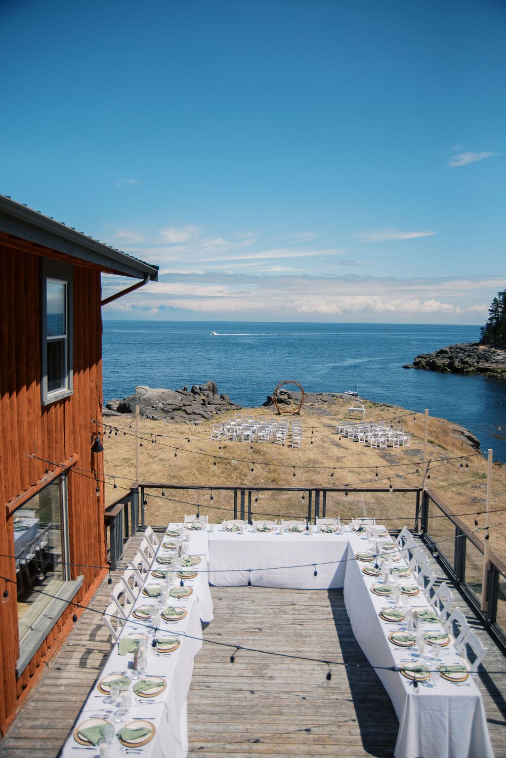 Elegant outdoor wedding reception tables set on a wooden deck overlooking a rocky coastline and blue ocean waters.