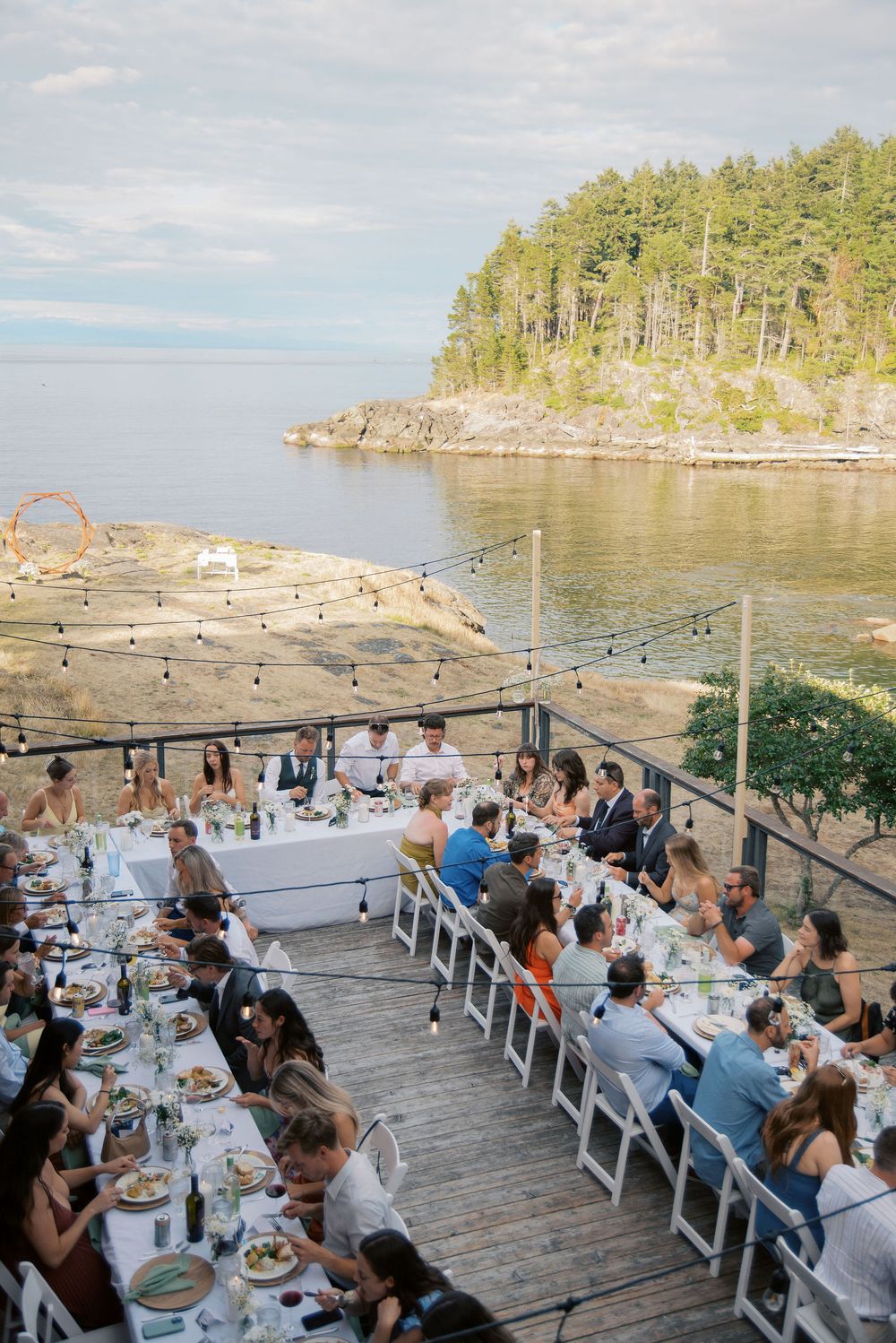Outdoor wedding reception on a waterfront deck with string lights, long dining tables, and guests enjoying a meal overlooking the ocean.