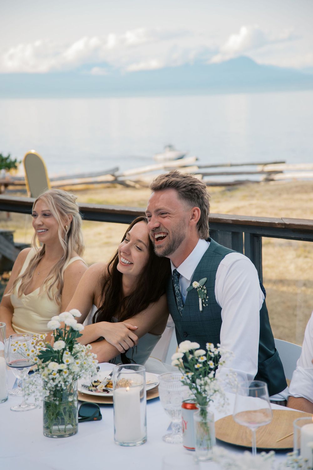 A laughing bride and groom sit together at an outdoor waterfront wedding reception table with floral centerpieces.