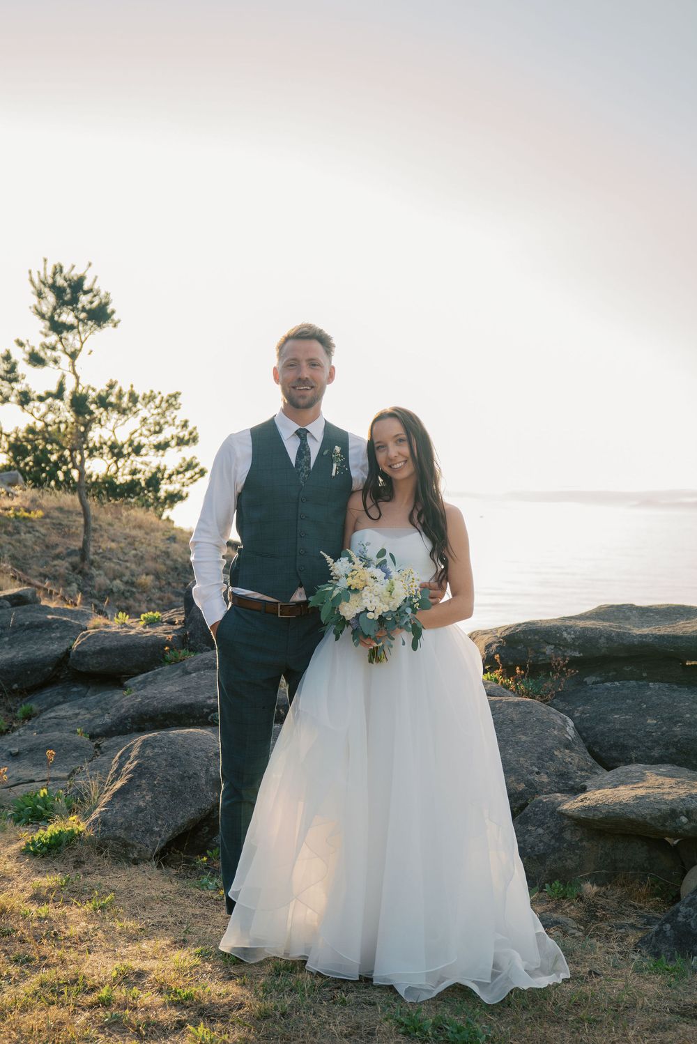 Bride and groom pose on rocky coastal bluff at sunset, she holds white bouquet in flowing gown, he wears dark vest.