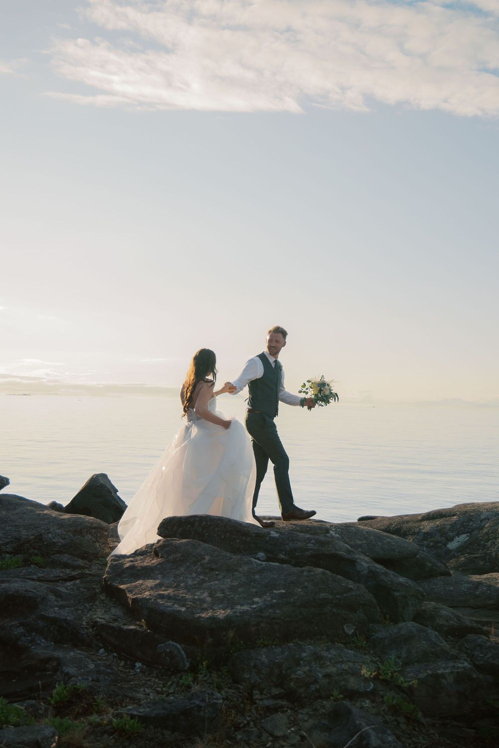 Bride and groom holding hands on rocky shoreline at sunset, groom holding bouquet, ocean backdrop.