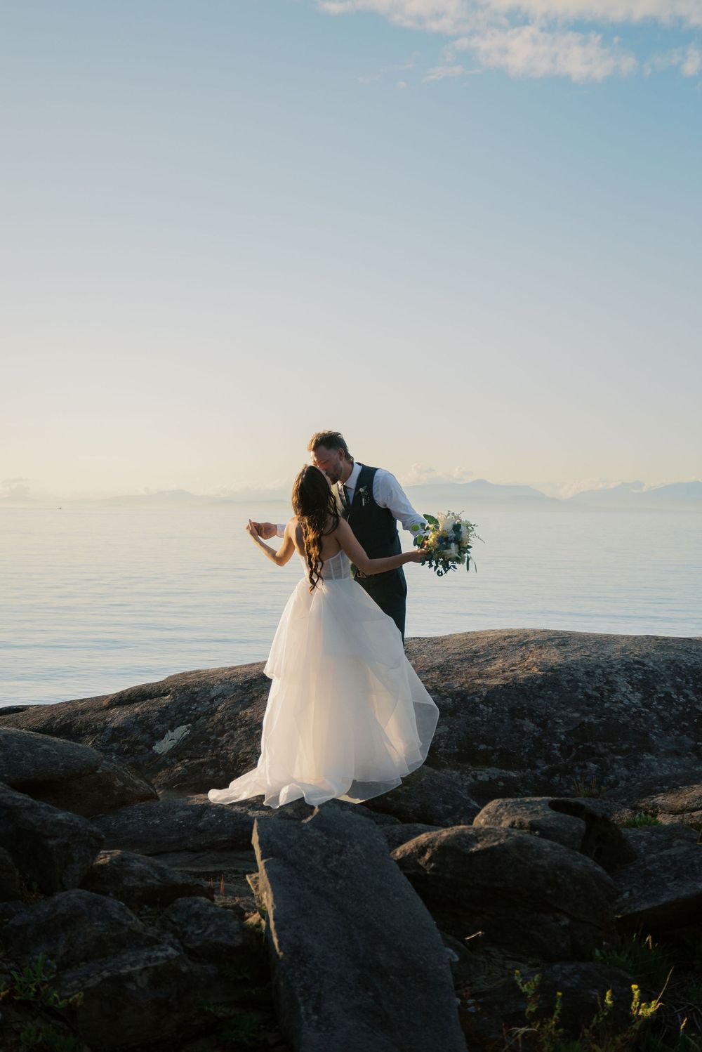 Bride and groom share a romantic kiss on rocky shoreline at sunset with calm ocean backdrop.