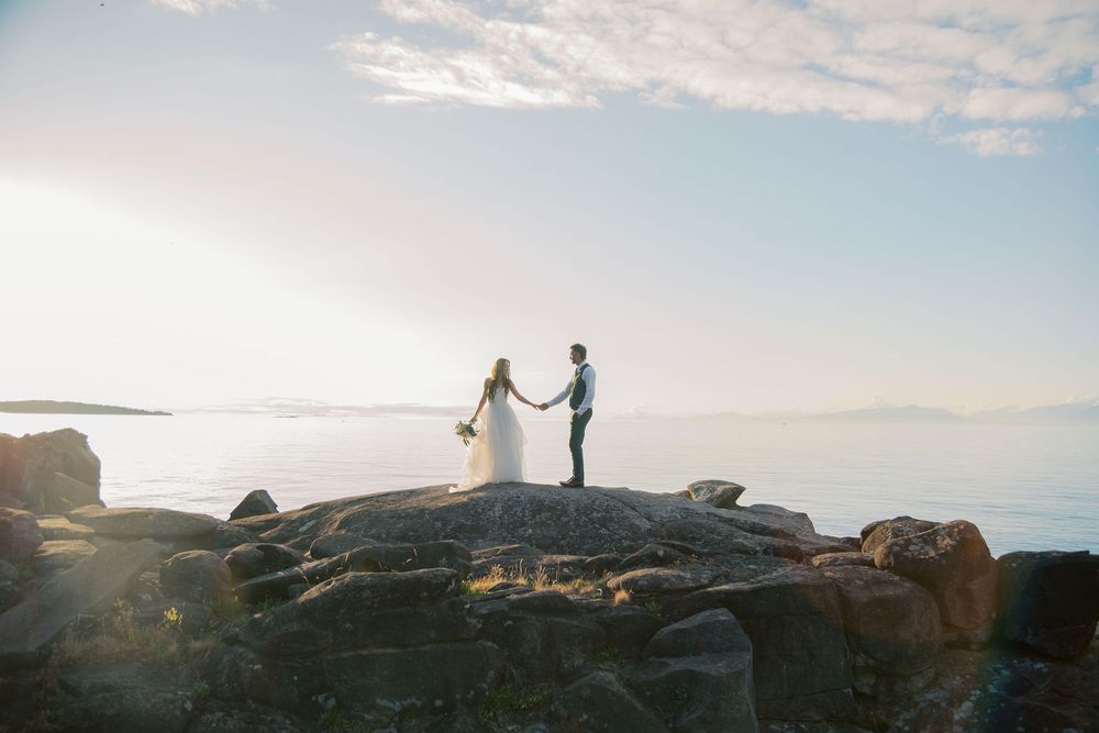 Bride and groom holding hands on rocky coastal cliffs with a stunning ocean backdrop during their outdoor wedding ceremony.