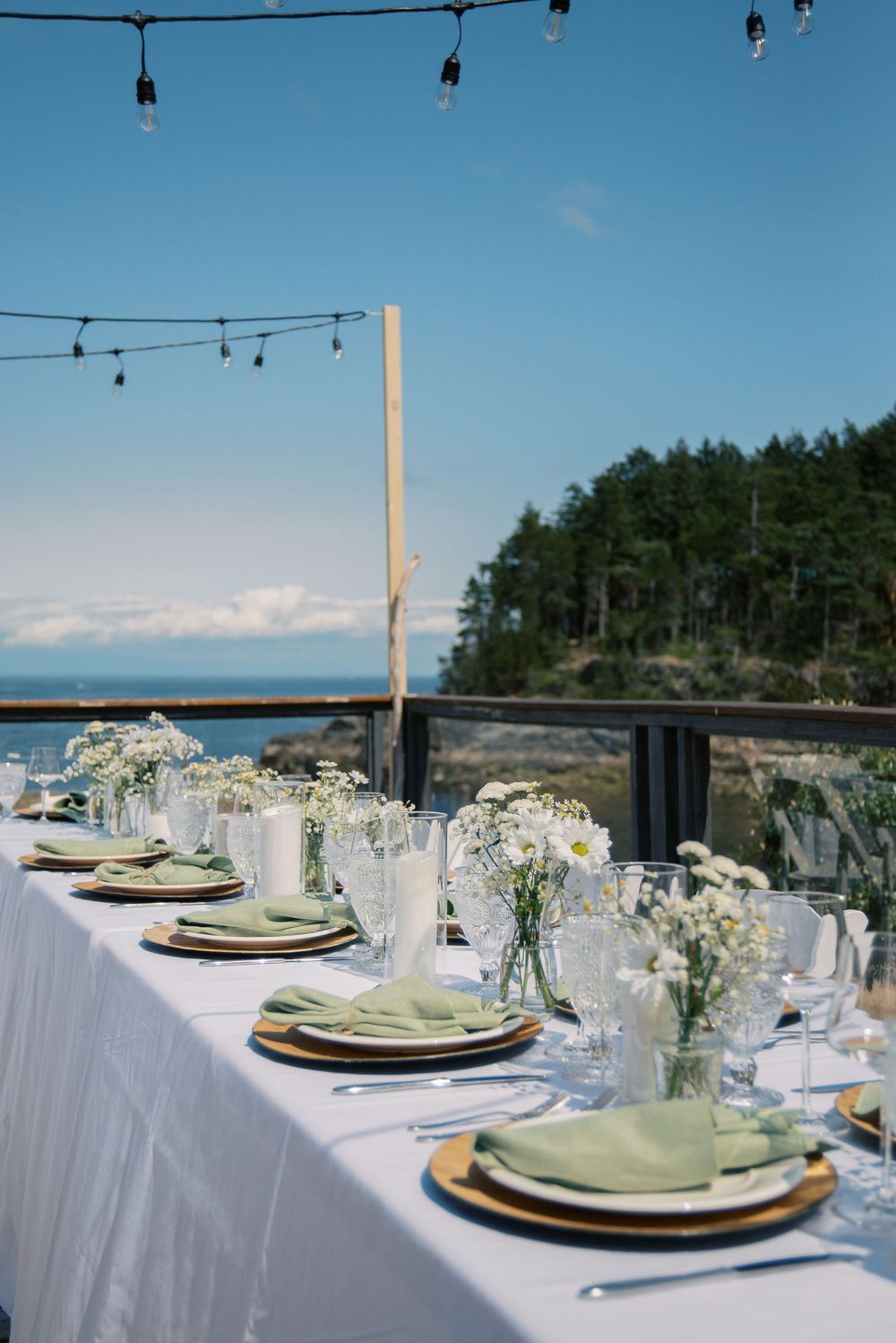 Elegant outdoor wedding reception table set with white linens, green napkins, wildflower centerpieces, and ocean views under string lights.