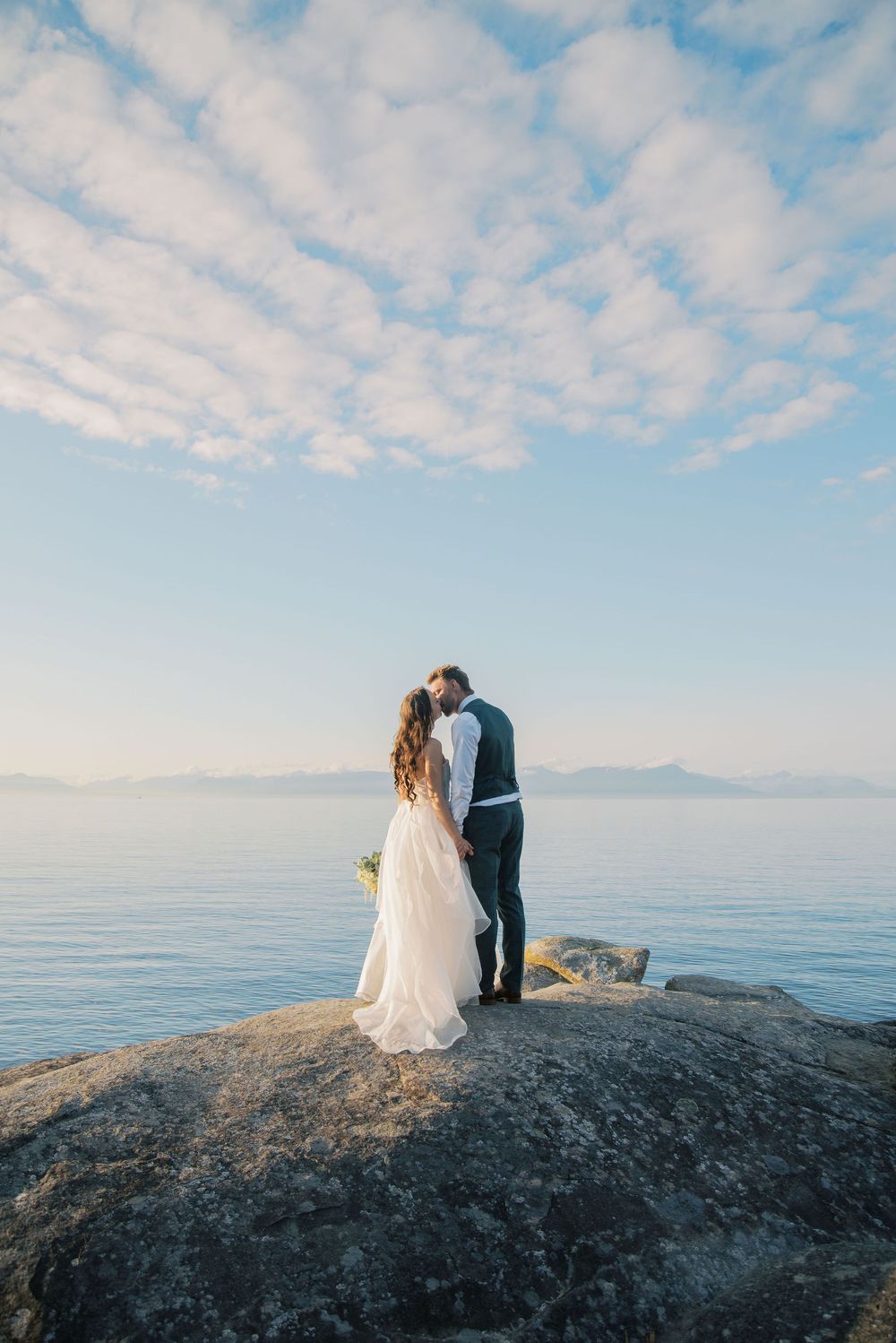Newlyweds embrace and kiss on rocky cliff overlooking serene ocean under dramatic cloudy sky.