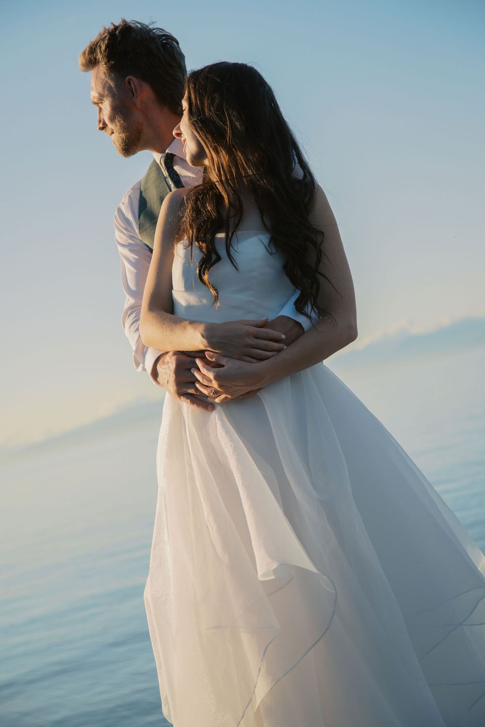 Romantic couple embracing by the ocean at sunset, bride in flowing white gown and groom in formal attire.