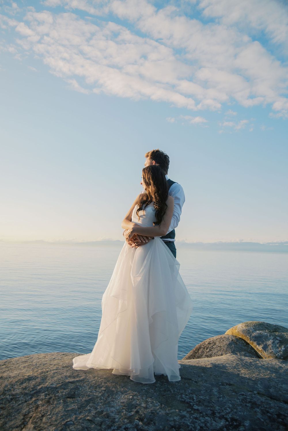 Bride and groom embracing on rocky shoreline, gazing at calm blue ocean under a partly cloudy sky.