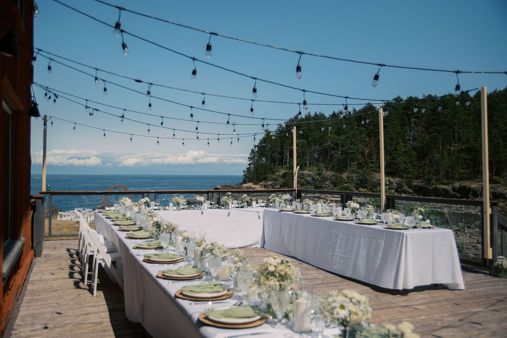 Elegant outdoor waterfront wedding reception with long white table, floral centerpieces, string lights, and ocean views.