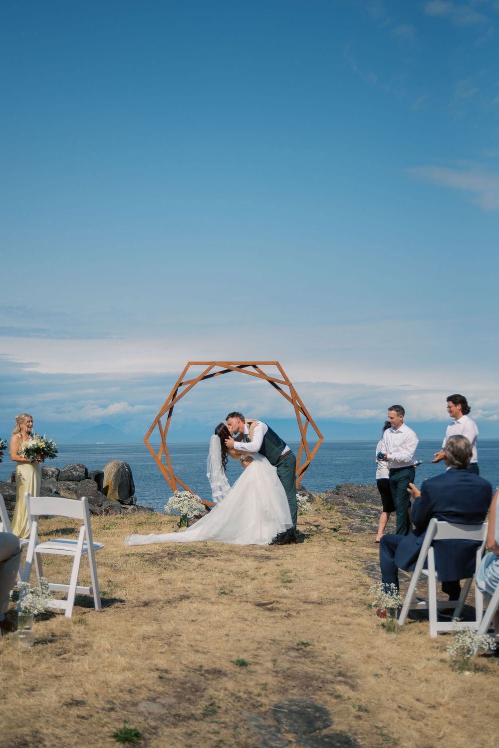 Couple sharing first kiss at outdoor coastal wedding ceremony under hexagonal arch with ocean backdrop.