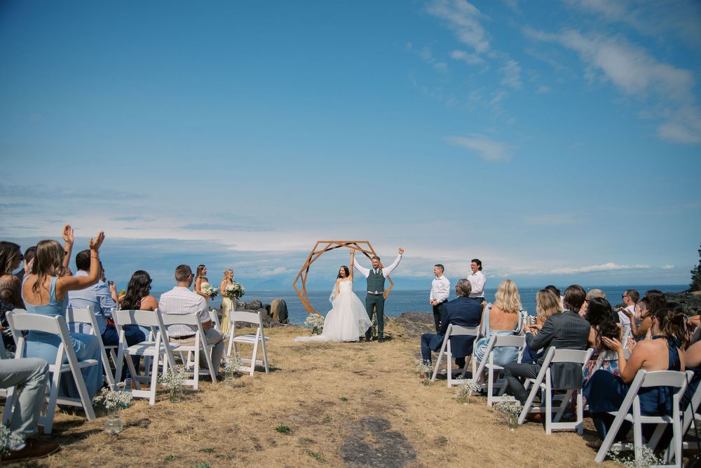 Outdoor beach wedding ceremony with couple under floral arch, guests seated on white chairs, ocean and blue sky backdrop.