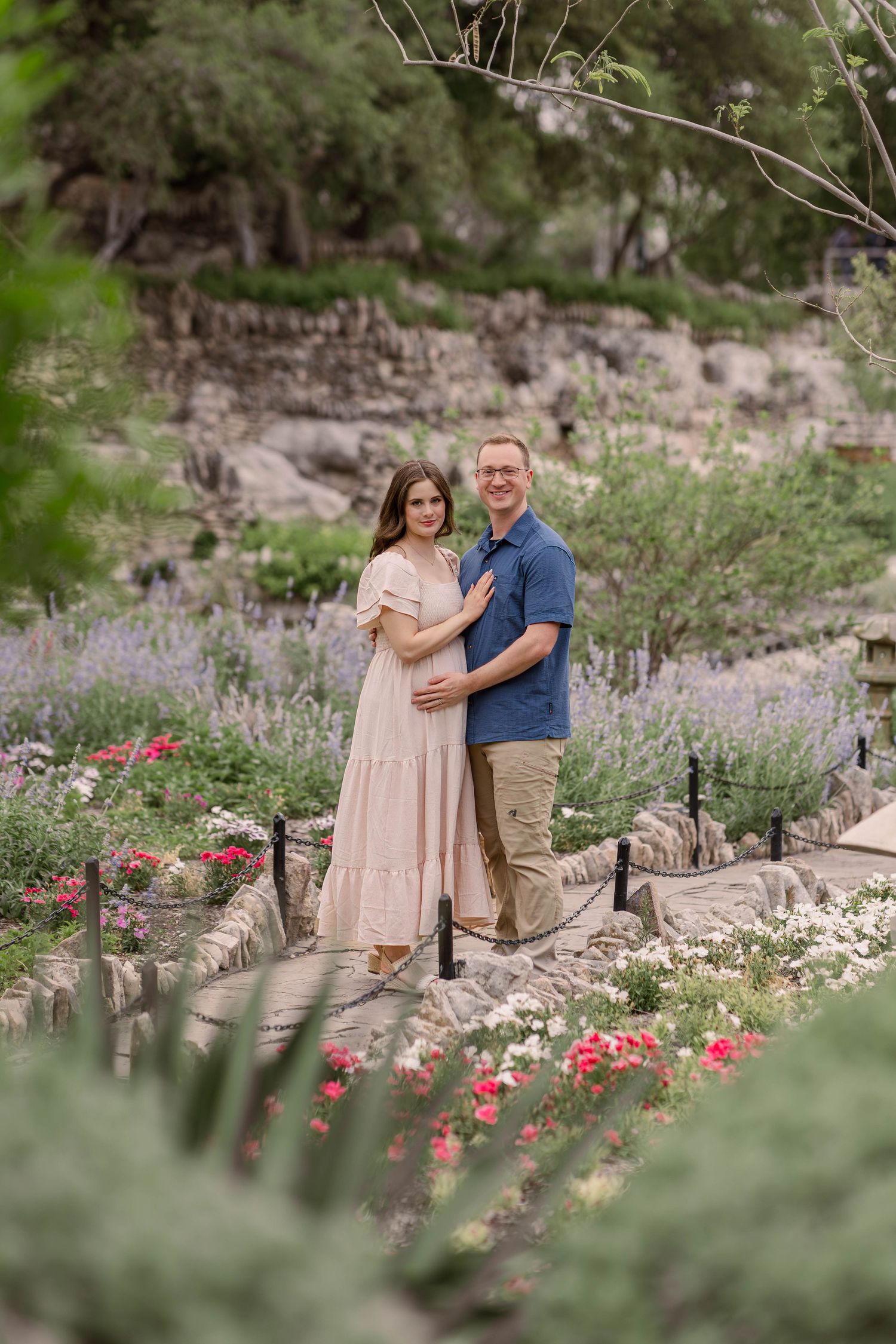 A couple poses in a flower garden, the woman in a pink dress cradling her baby bump, the man in a blue shirt embracing her.