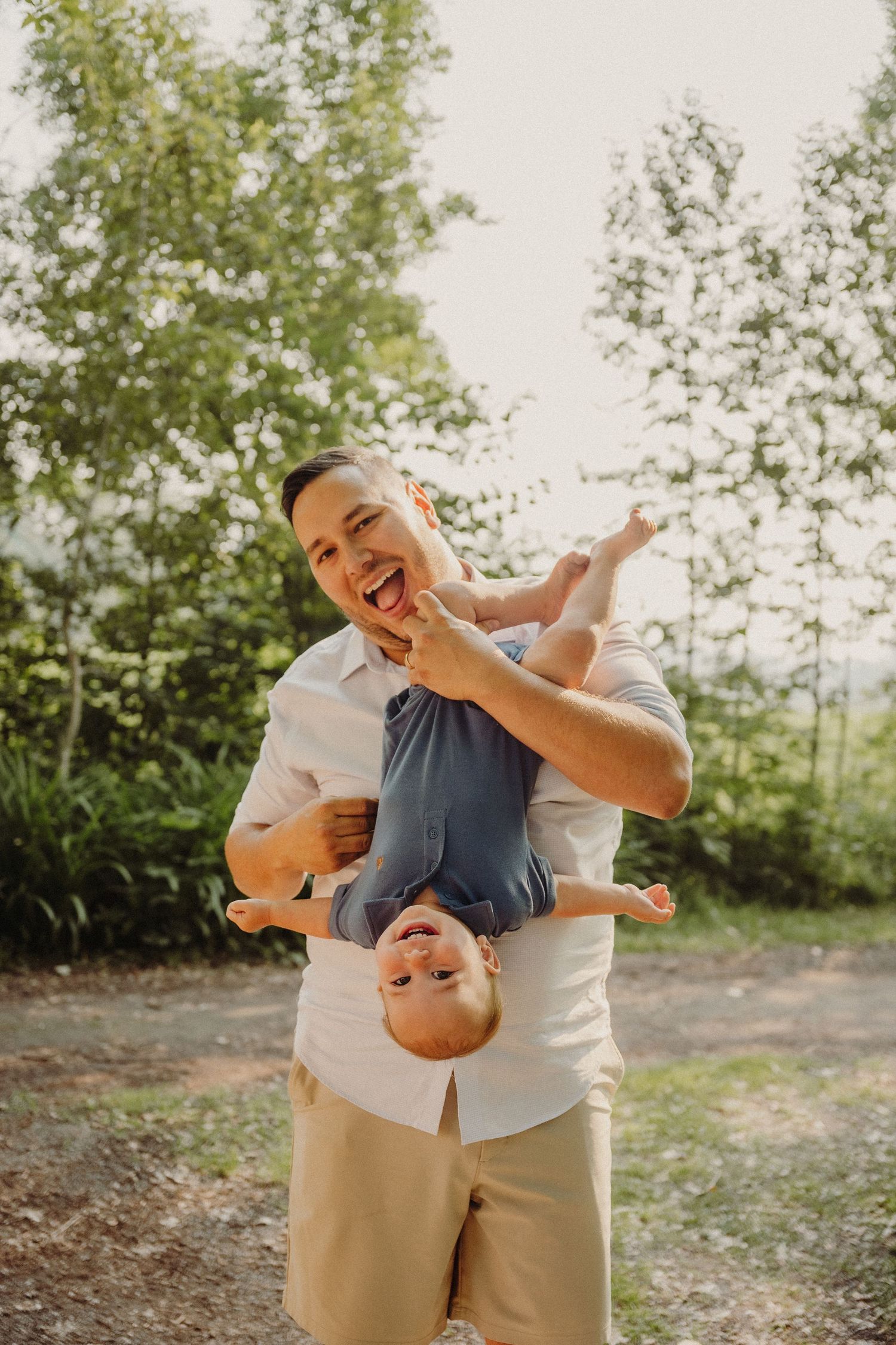 A laughing man playfully holds a young child upside down outdoors, both smiling joyfully in a lush green setting.