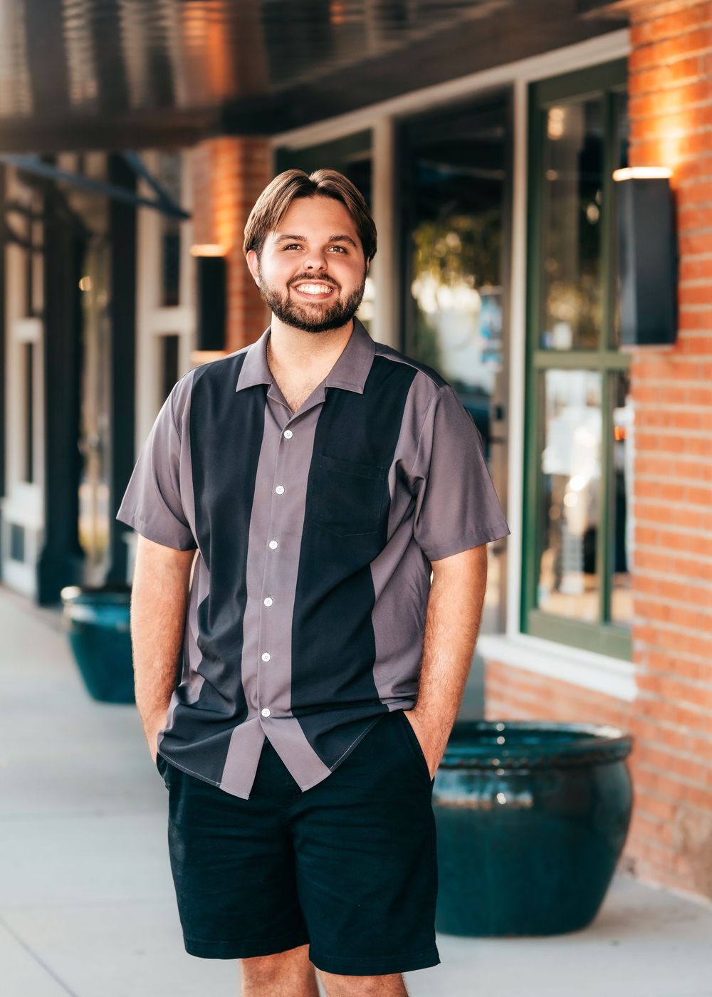 Smiling young man with beard wearing gray and black bowling shirt and black shorts standing outside brick building.