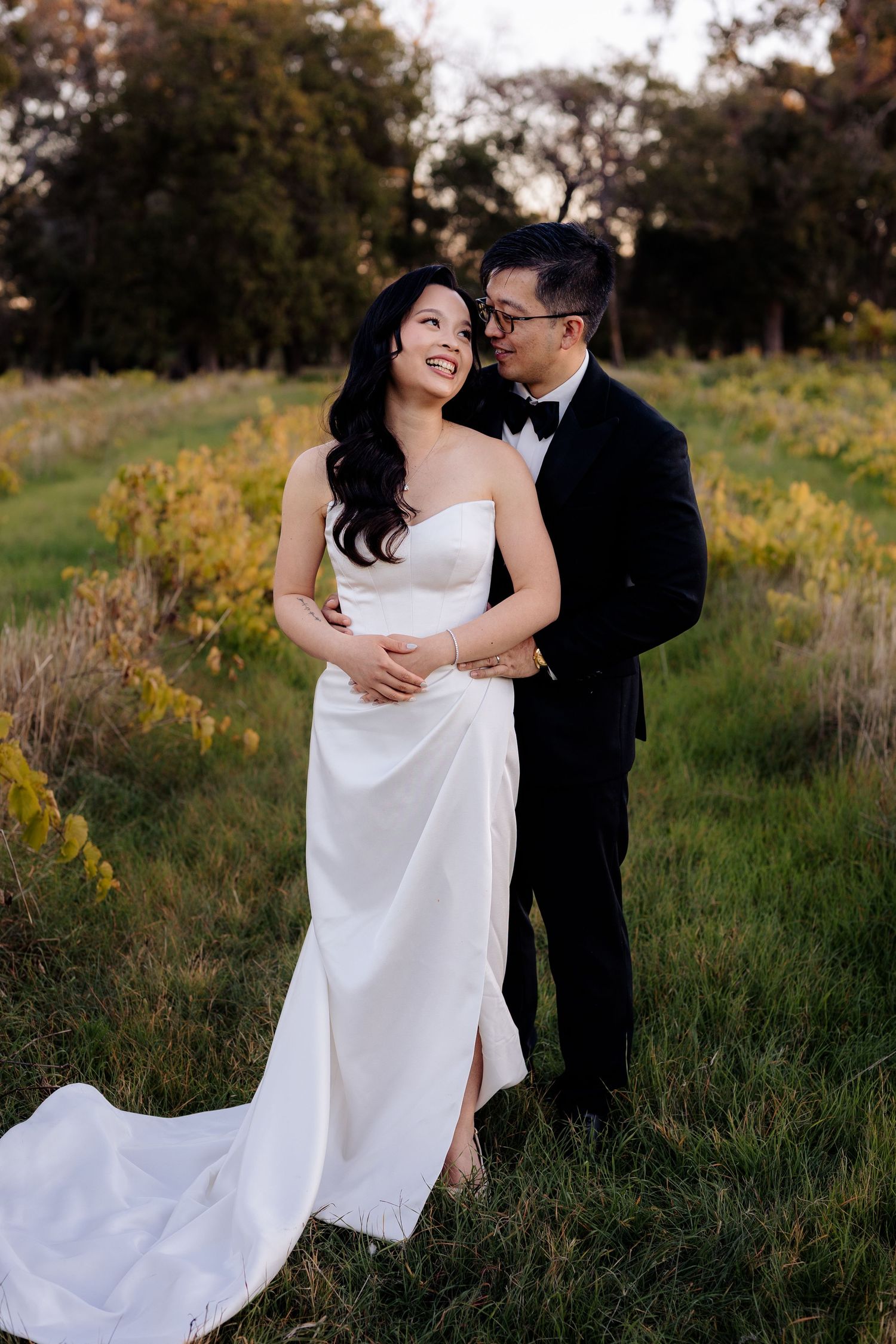 Bride and groom embracing in a green field at golden hour, wearing a white gown and black tuxedo.