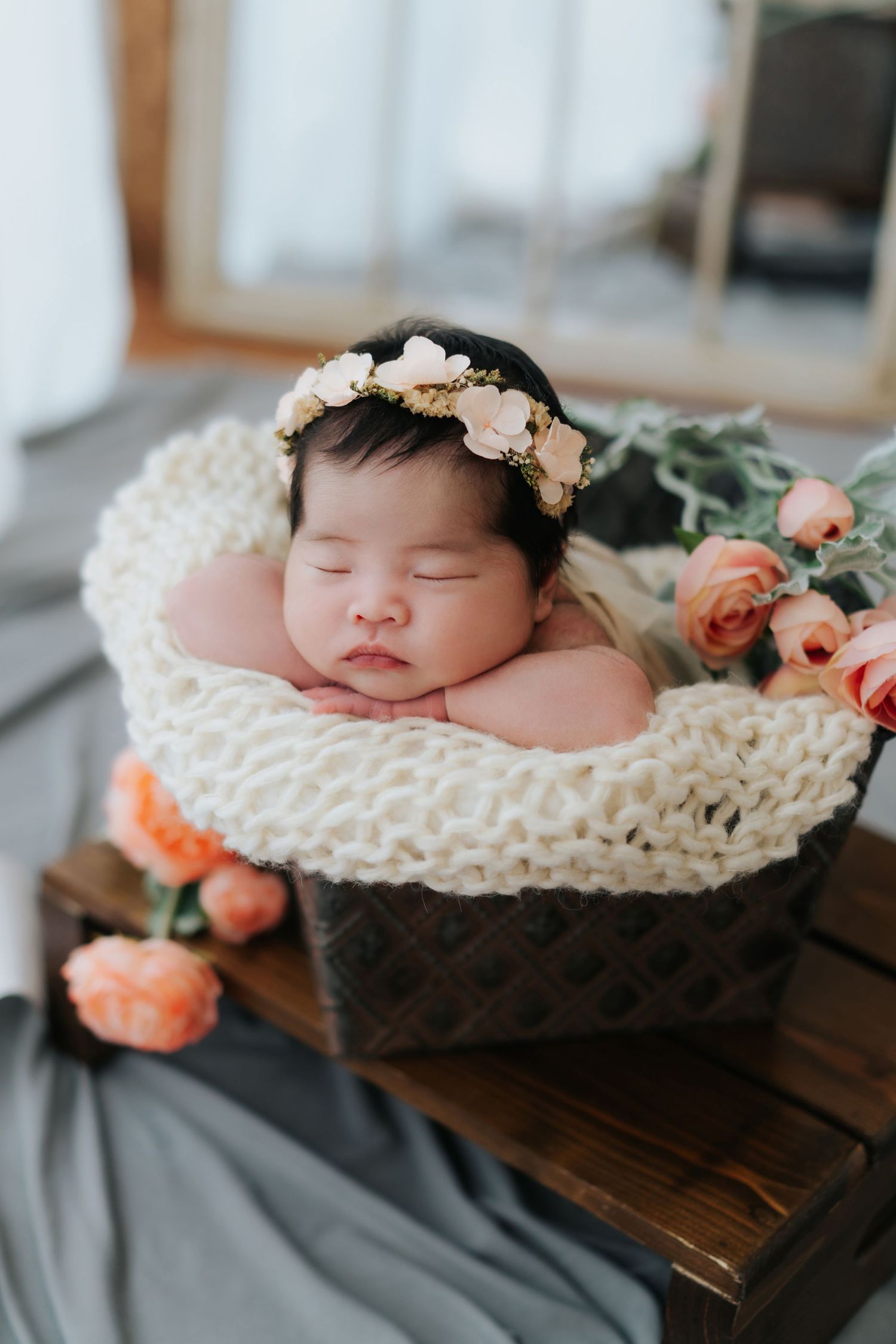 Sleeping newborn baby in a woven basket with a floral headband, surrounded by peach roses.
