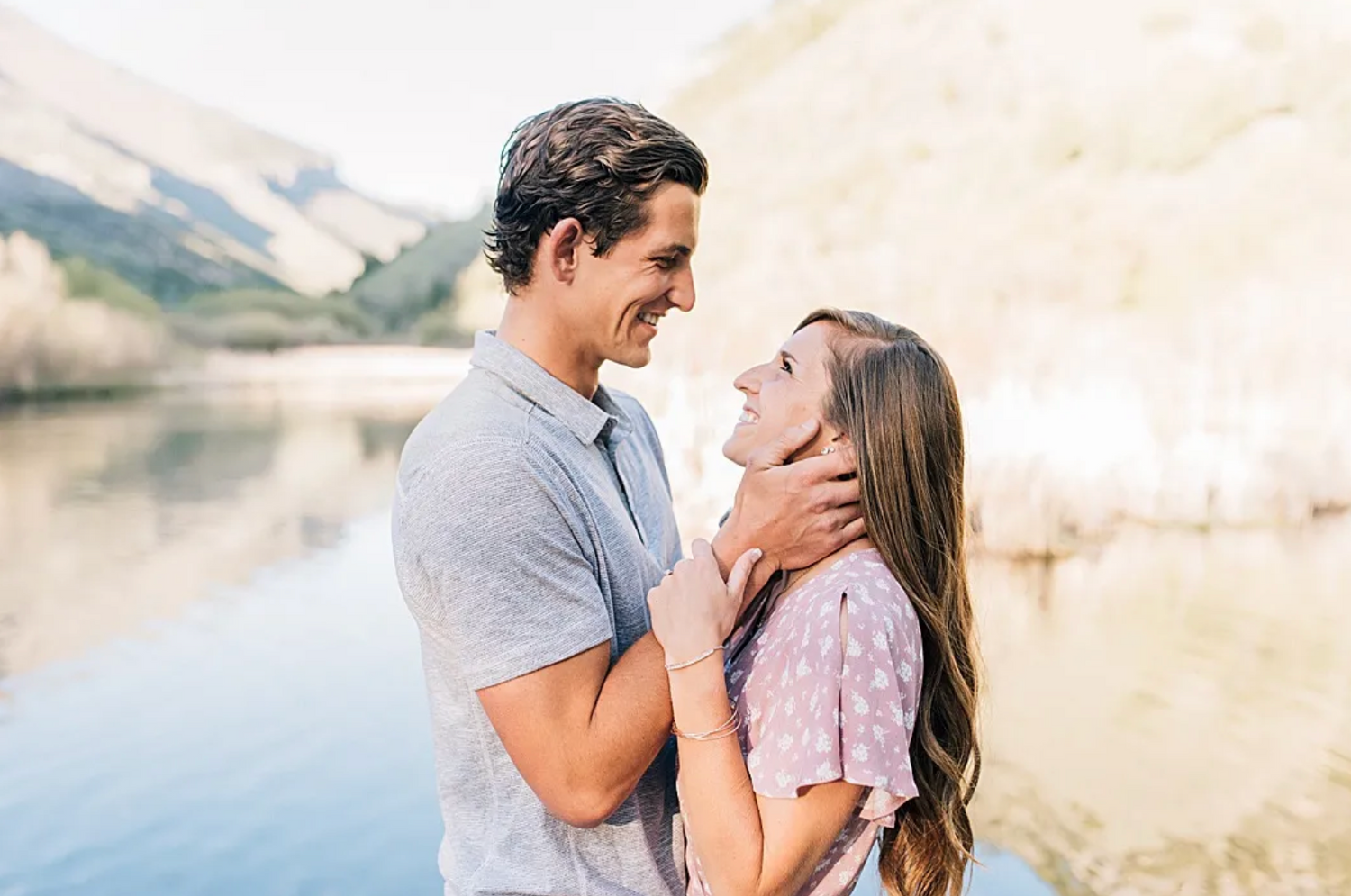 A romantic couple embraces by a serene mountain lake with soft sunlight and natural scenery in the background.