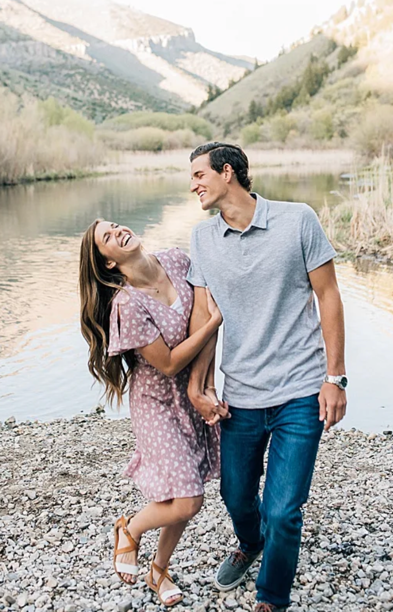 A couple shares a joyful moment while holding hands along a rocky shore with mountains and a lake in the background.