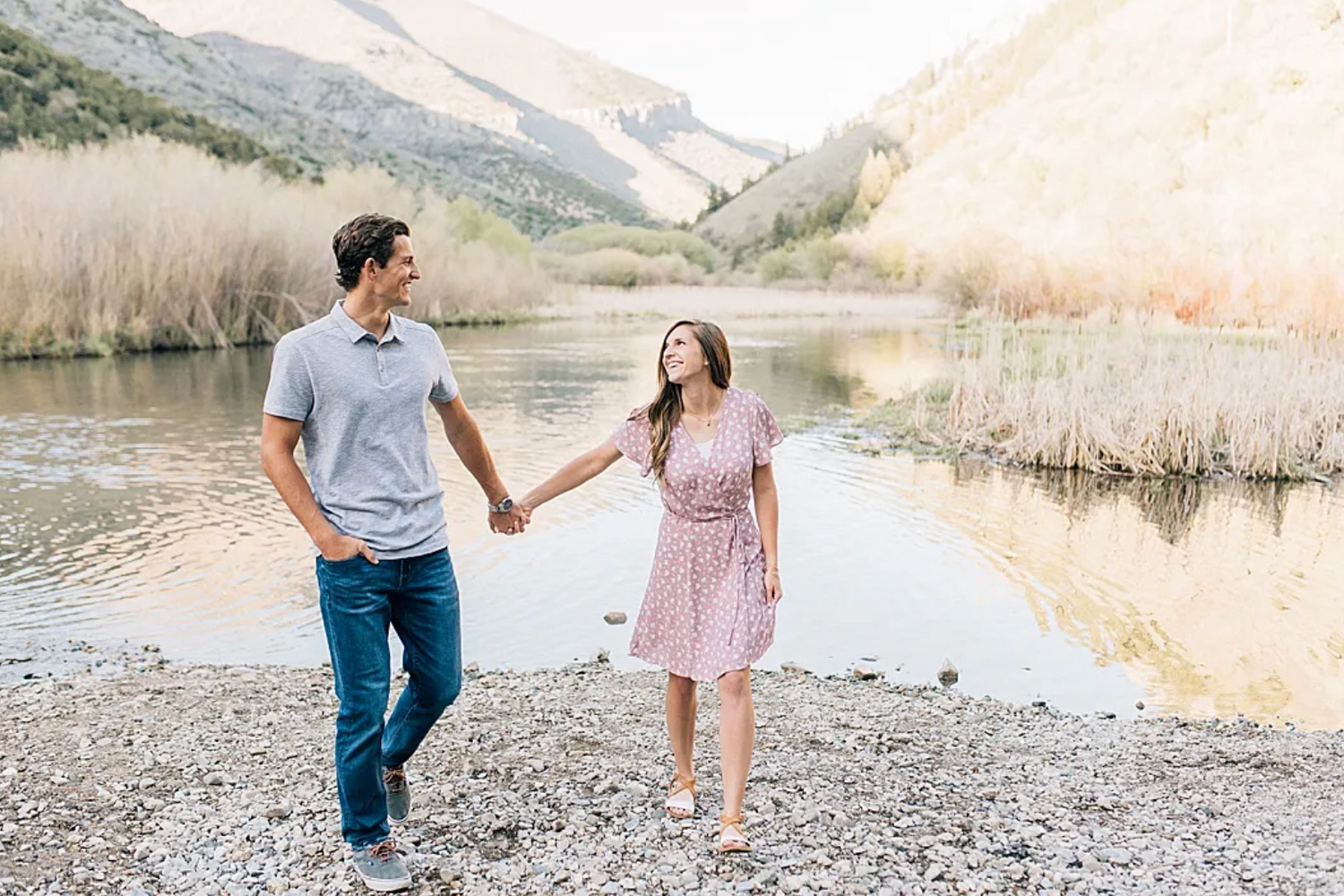 A couple walks hand in hand by a serene mountain lake surrounded by tall reeds and peaks during sunset.