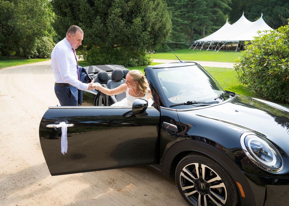 A bride in a white dress sits in a black Mini Cooper convertible as a man holds the door open at a wedding venue.
