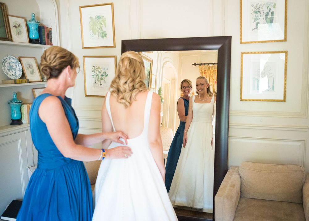 Bridesmaid in blue dress helps bride with wedding gown while reflection shows in large floor mirror indoors.