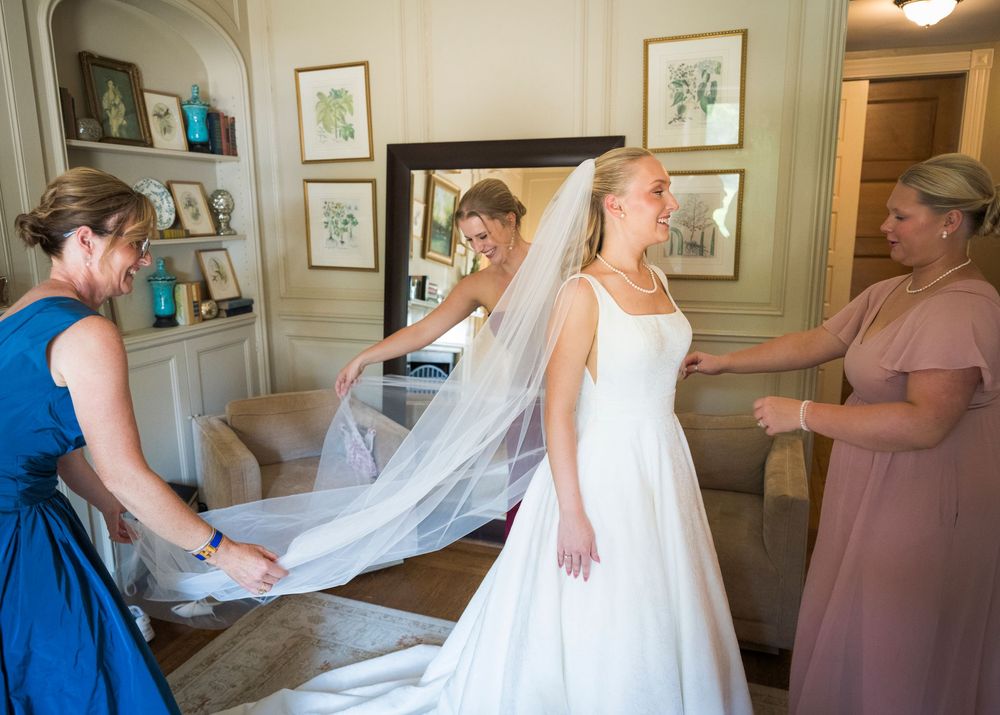 Bride in white gown and veil being helped by bridesmaids in blue and pink dresses in elegant room.