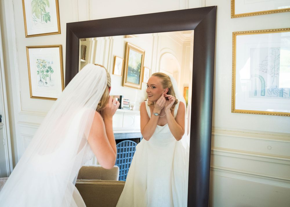 Bride in white gown and veil adjusting earrings while looking in a large ornate mirror before her wedding.