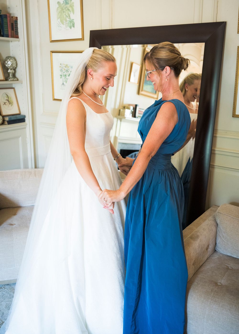 Bride in white gown and veil holds hands with woman in blue dress smiling beside large mirror.