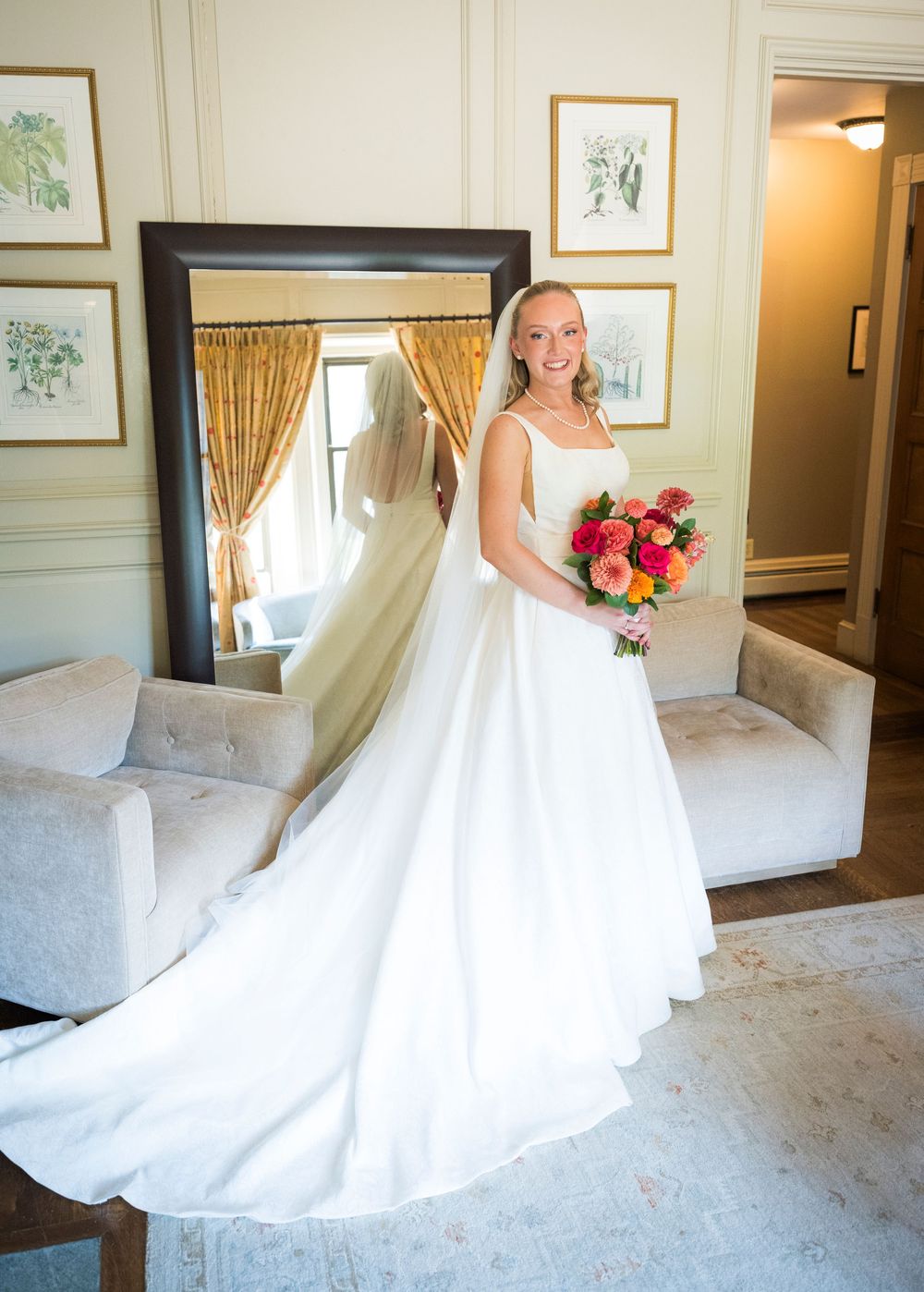 Bride in elegant white gown holding colorful bouquet stands before large mirror in decorated bridal suite.