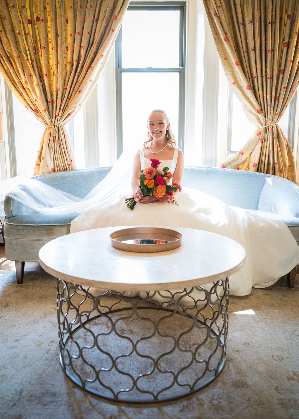Smiling bride in white gown holding colorful bouquet seated on blue sofa near window with gold curtains.