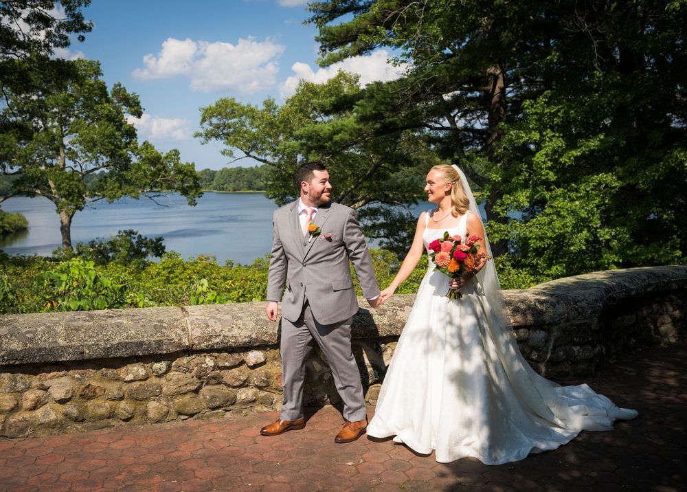 Bride and groom walk hand in hand on a brick terrace overlooking a serene lake surrounded by lush trees.